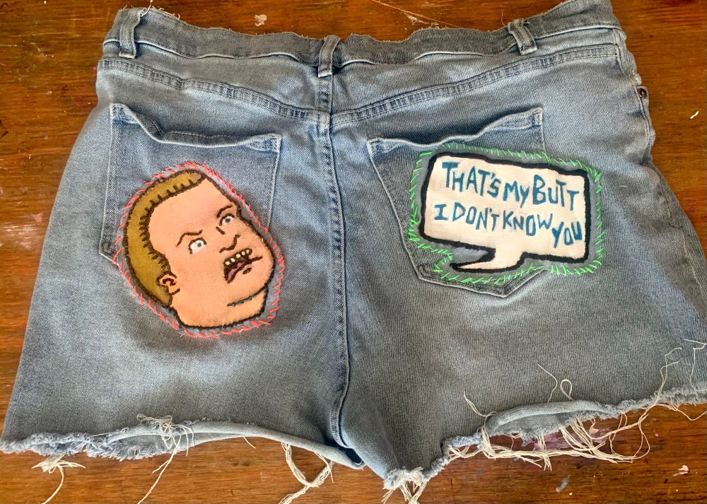 A pair of cutoff shorts with hand stitched patches on the back featuring Bobby Hill shouting “That’s my butt I don’t know you”