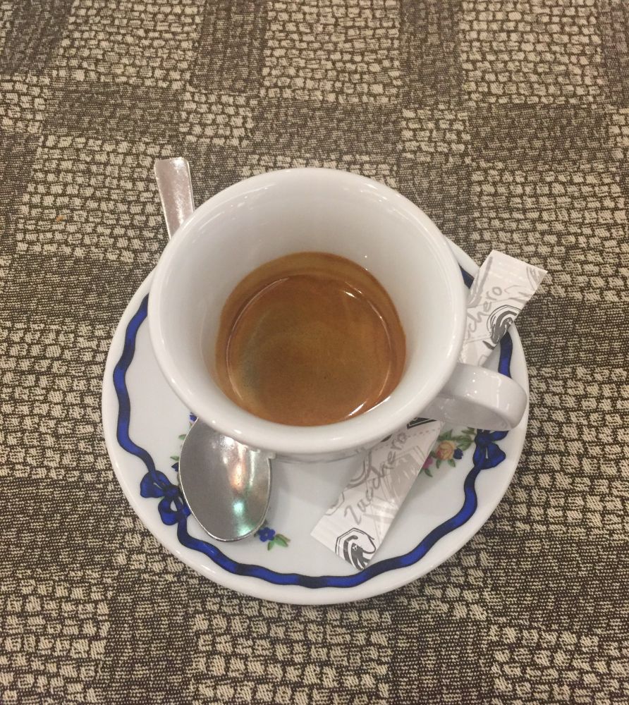 A tiny cup with an even tinier shot of espresso in it. It’s sat on a table top with a mosaic pattern. The saucer is white with a blue ribbon pattern. There’s a teaspoon and a sugar packet on the saucer. This is Izzy’s coffee order in Rome.