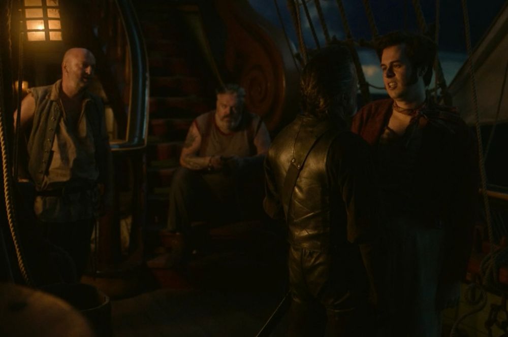 A scene from episode five, series one of Our Flag Means Death. It’s the deck of the Revenge at night. Pete (Matt Maher) stands on the left, Wee John (Kristian Nairn) sits on the stairs and to the right, Lucius Spriggs (Nathan Foad) is stood facing Izzy Hands (Con O’Neill). They’re having a bit of a stand off, which Pete is in prime position to enjoy!