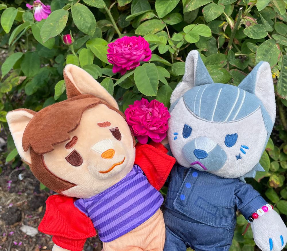 A Lucius Spriggs cat plushie and an Izzy Hands dog plushie, cuddling in front of a bush of deep pink roses. 