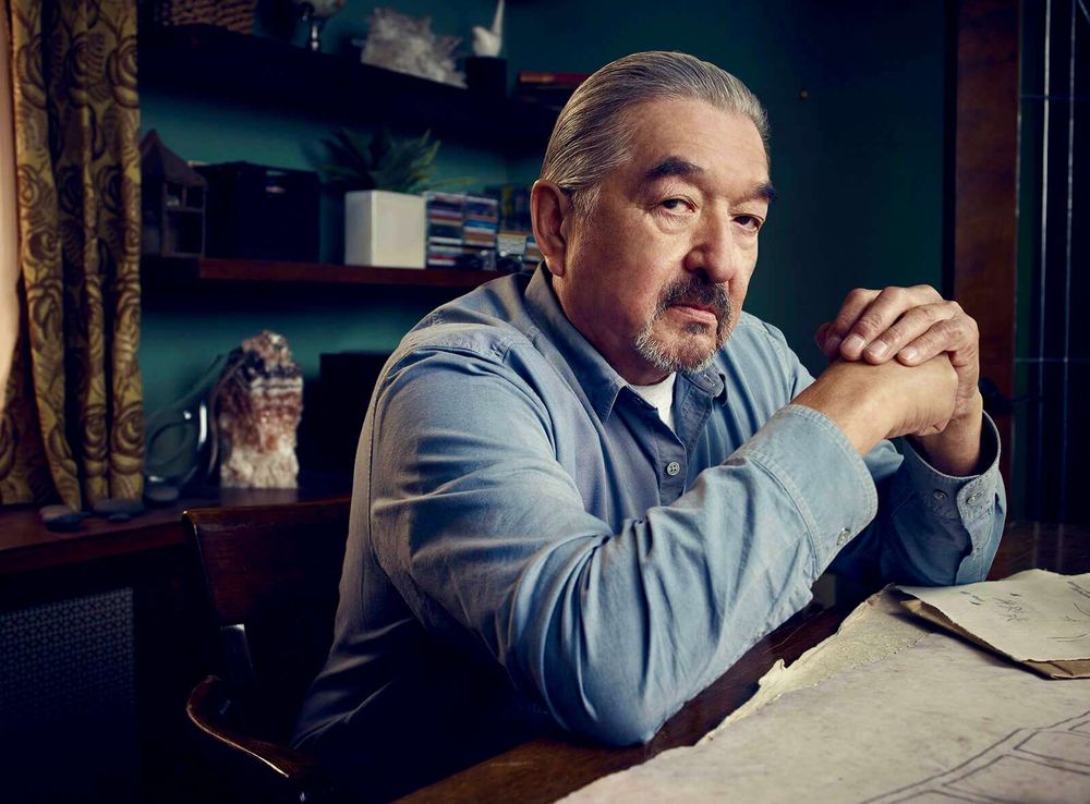A colour photo of actor Graham Greene loking at the camera. He is wearing a blue shirt, sitting at a desk.