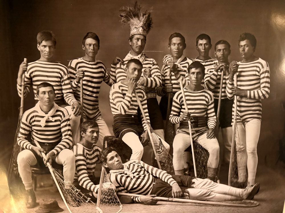 A William Notman Studio photograph of the Kahnawà:ke lacrosse team that toured Great Britain in 1876. A black and white team photo: the uniforms were red and white striped jerseys with blue shorts. 