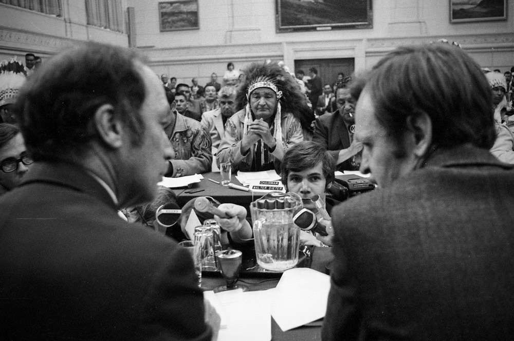 A photograph of Walter Deiter, in a headdress, presenting to PM Pierre Trudeau and Indian Affairs Minister Jean Chretien, whose backs are to the camera.