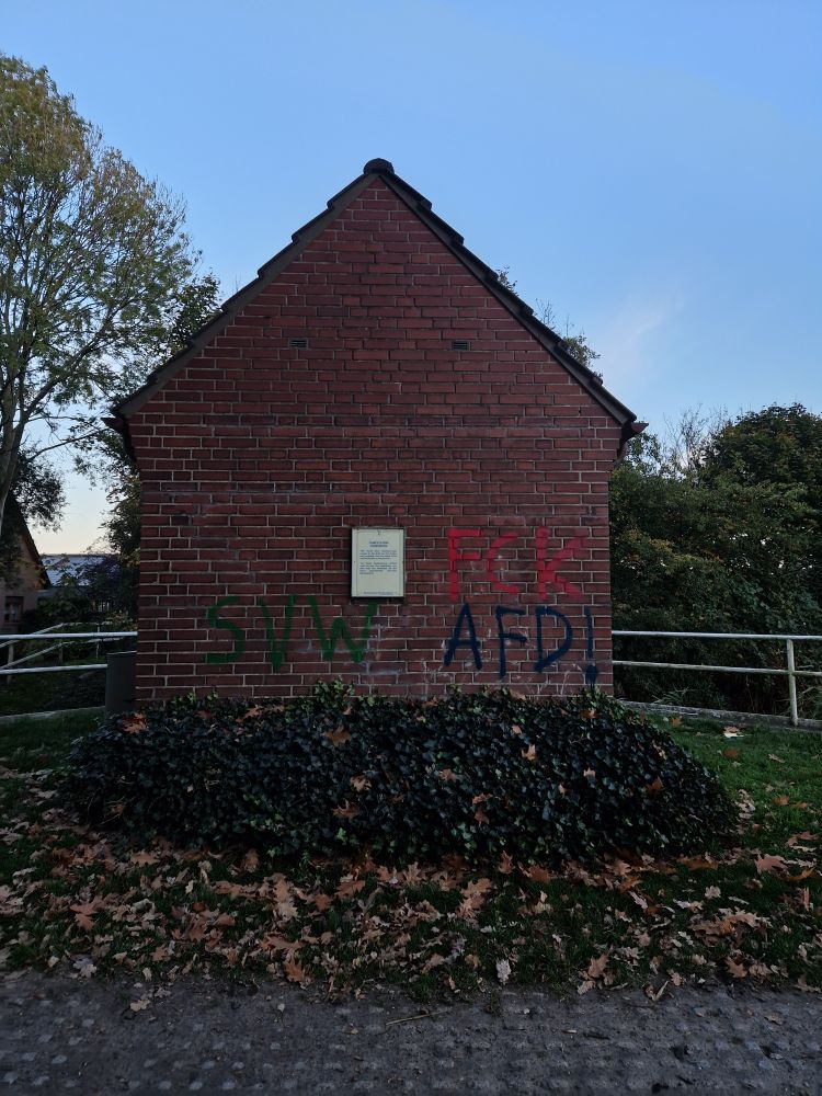 Giebelseite eines kleinen Häuschens aus rotem Klinker und schwarzem Dach ohne Fenster oder Tür. Rechts und links ein eine weißes Geländer aus Stahlrohr, dahinter grüne Bäume und Sträucher, blauer Himmel. Vorm Häuschen Efeu und Gitterpflastersteine.
Das Häuschen ist besprüht mit "SVW" und "FCK AFD!" 
