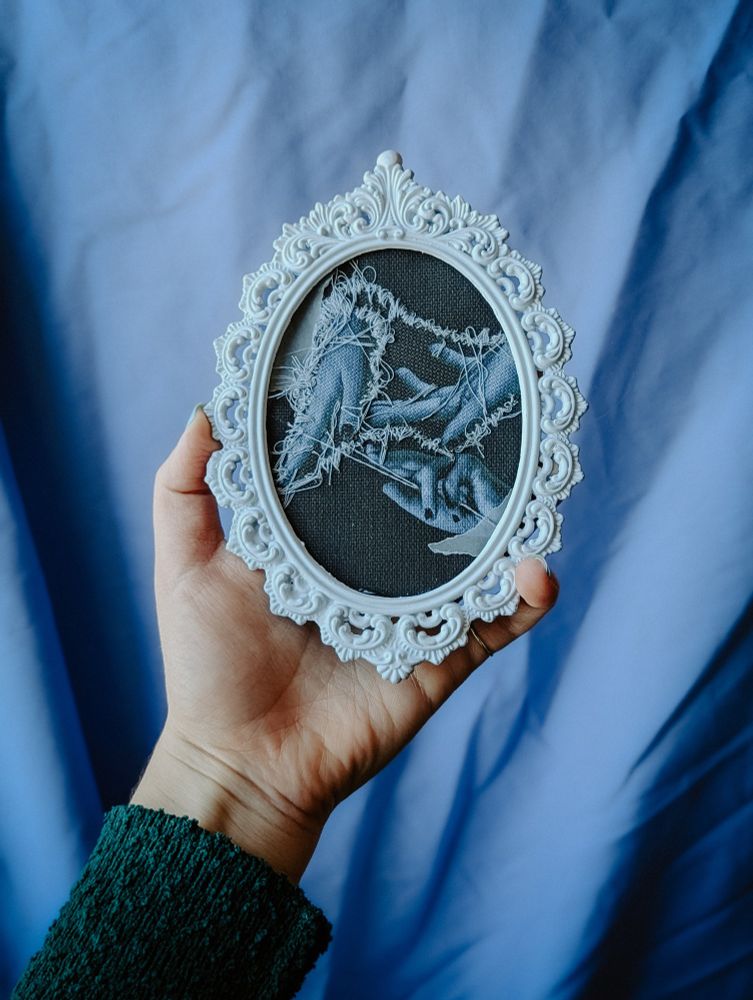 Photo of Annie's hand holding her Sweetest Little Thing donation. It's a vintage baroque-esque white metal frame a bit bigger than her hand. Inside 3 blue toned hands are intertwined with baby blue thread. It's messy and imperfect. You can see the emerald green sleeve of Annie's cardigan and the backdrop is a blue textile.