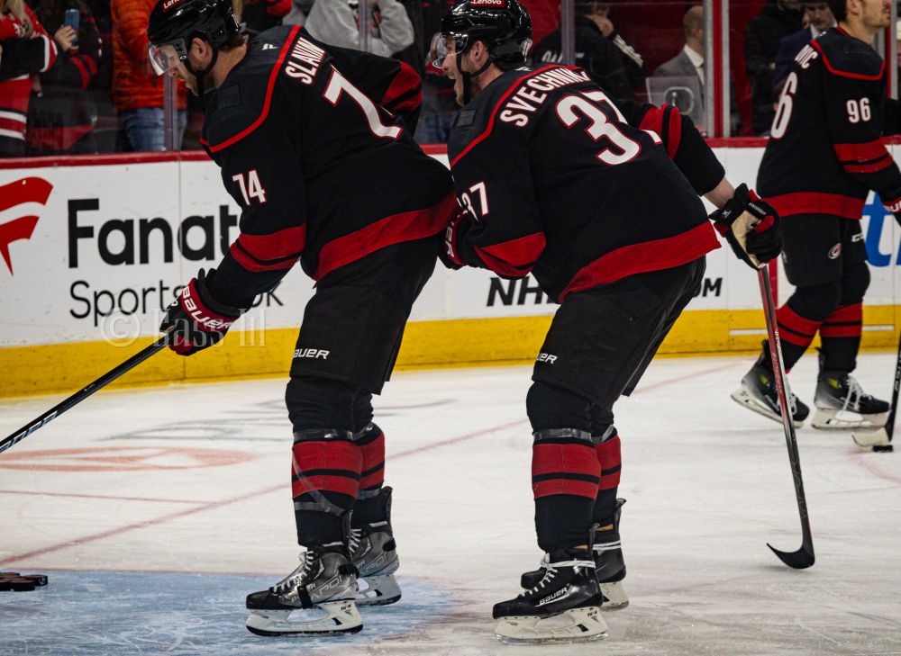 Jaccob Slavin fishing some pucks out of the net during warm ups while Andrei Svechnikov tugs on his jersey from behind and laughs
