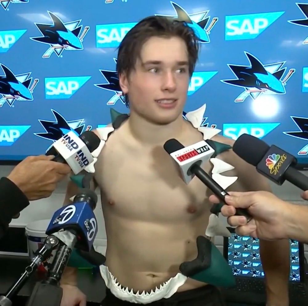 Macklin Celebrini wearing a giant necklace completely made of shark teeth that hangs down to his pants. Some of the teeth are painted in sharks colors and the bottom part is an entire small shark jaw. He is also shirtless and being interviewed post game so there are several microphones in his face 