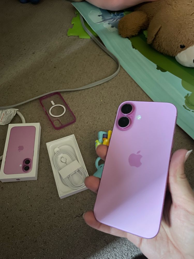 On the right, a hand is holding up a pink IPhone 16 with the packaging and phone case scattered on the floor to the left.