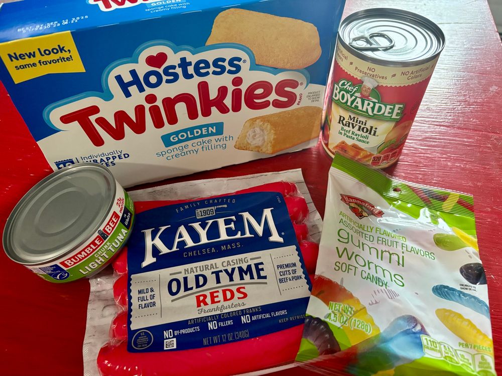 A package of twinkies, tuna in a can, red hot dogs, gummy worms, and mini raviolis