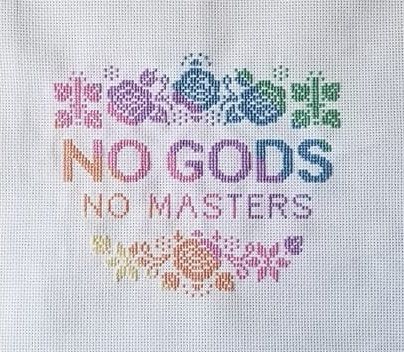 text that says no gods no masters surrounded by floral and upside down cross motif done in a rainbow pastel gradient as cross stitch embroidery on white aida cloth