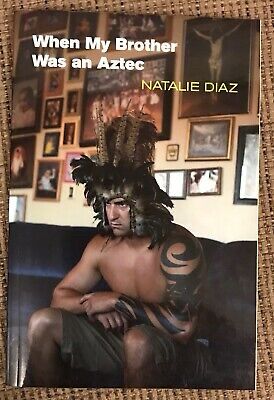 The image shows the cover of the book "When My Brother Was an Aztec" by Natalie Diaz. The cover features a muscular man sitting on a dark couch, adorned with a striking feathered headdress that adds a dramatic flair to the composition. His body is partially visible, showcasing tribal tattoos on his arm, which contribute to the cultural theme of the book. Behind him, a wall is filled with an array of framed photographs, creating a personal and intimate backdrop that suggests a connection to family and heritage. The title of the book is prominently displayed at the top in bold white letters, while the author's name, Natalie Diaz, is positioned below in a bright yellow font, making it stand out against the darker tones of the image. The overall aesthetic combines elements of strength and cultural identity, inviting readers to explore the themes within the book. The warm color palette enhances the inviting feel of the cover, making it visually appealing and thought-provoking. - from @coolhand.bsky.social alt text app. 