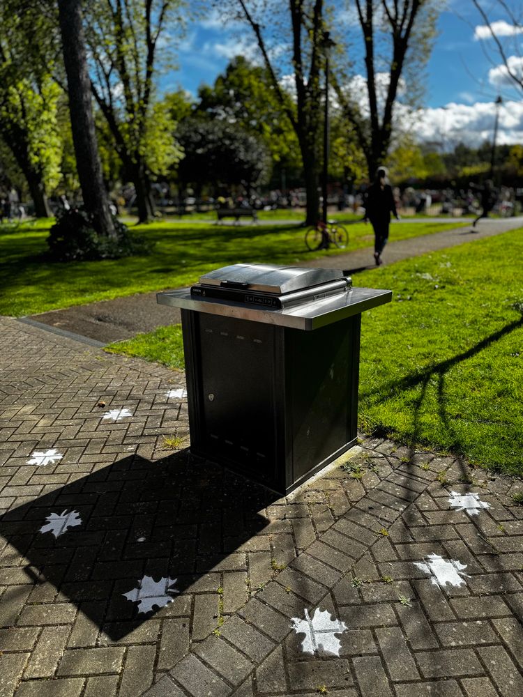 Photo of a park setting. It's a sunny day with some trees and people blurred in the background. In the foreground is a cooking appliance with a hood. Around it is a ring of stencil leaf markings. 