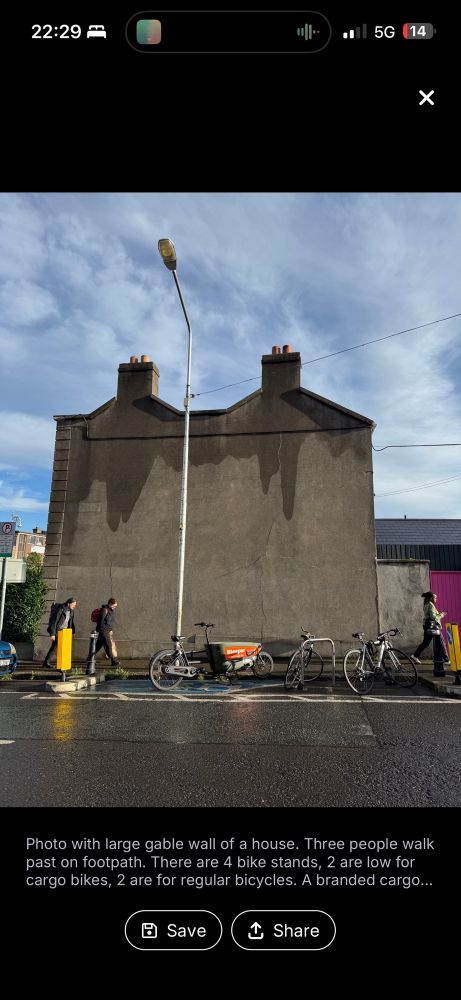 Photo with large gable wall of a house. Three people walk past on footpath. There are 4 bike stands, 2 are low for cargo bikes, 2 are for regular bicycles. A branded cargo is locked to one of the stands. 