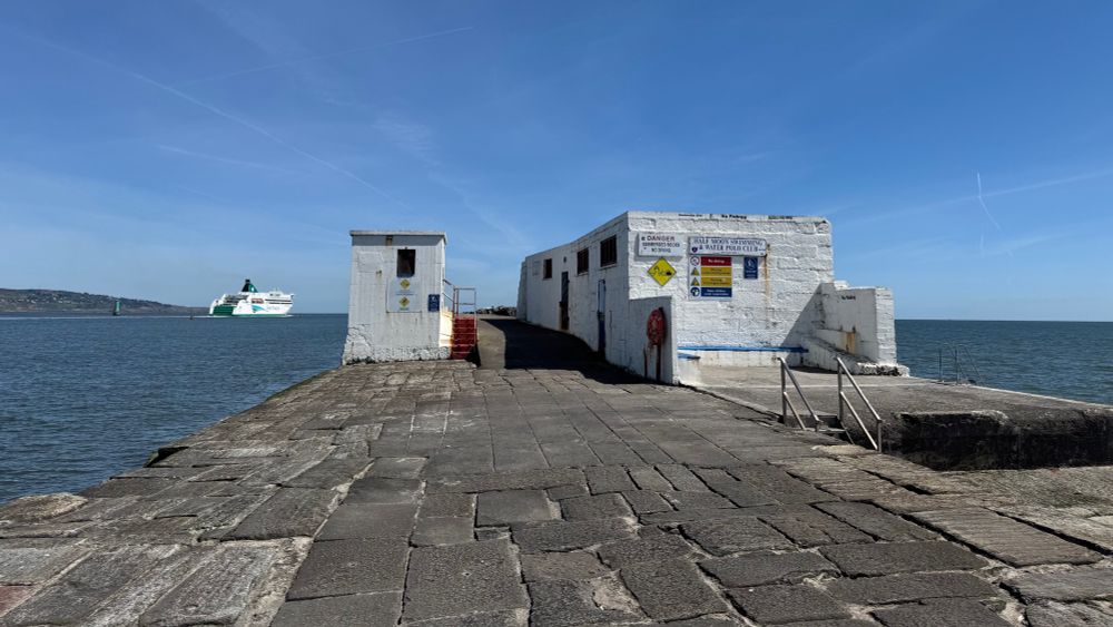 Photo of the South Wall at the point of the Half Moon Club. Two white buildings - either side of the causeway - and blue sky, make it feel like Greece. Howth Head is and a ferry leaving port are distant left. 