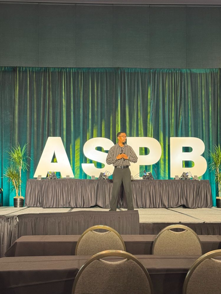 A speaker stands in front of an audience. Behind him are green curtains, and big block white letters that say 'ASPB.'