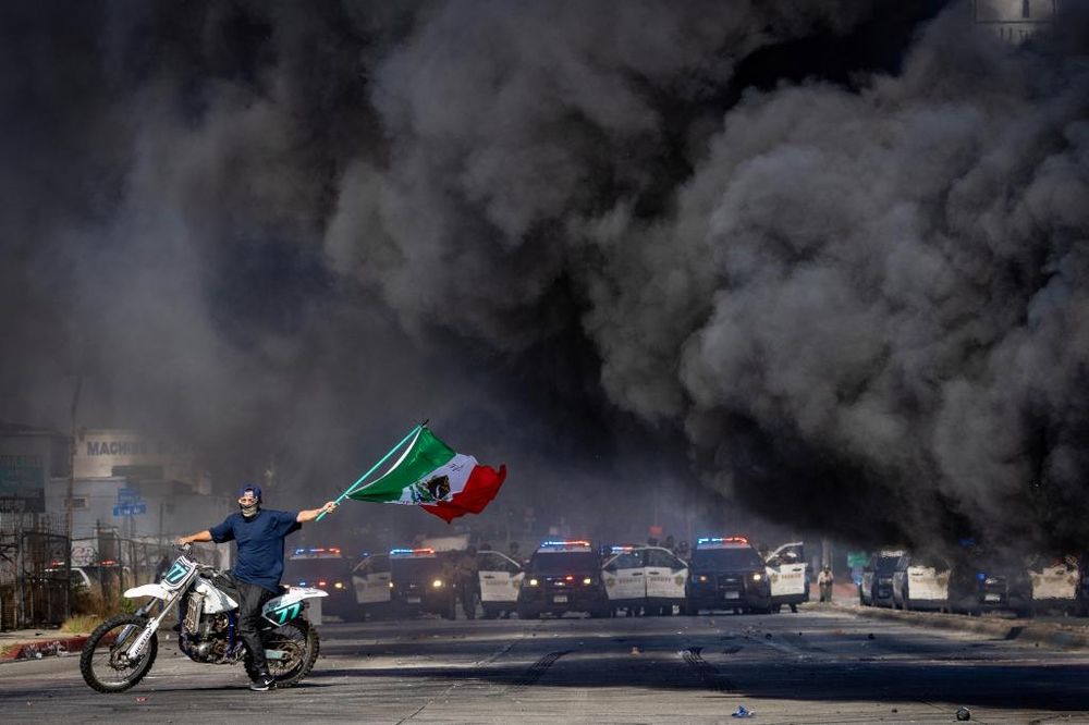 the coolest dude ever, on a dirt bike with a skull half-mask, waves a Mexican flag in Los Angeles in front of a line of police cars as billowing black smoke blows across the frame