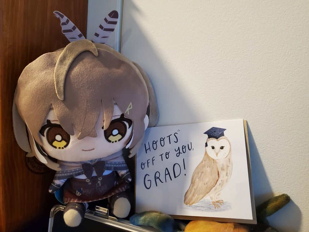 Nanashi Mumei plush sitting next to a card. The card has a picture of an owl with a graduation cap and text that says "Hoots off to you, grad!"