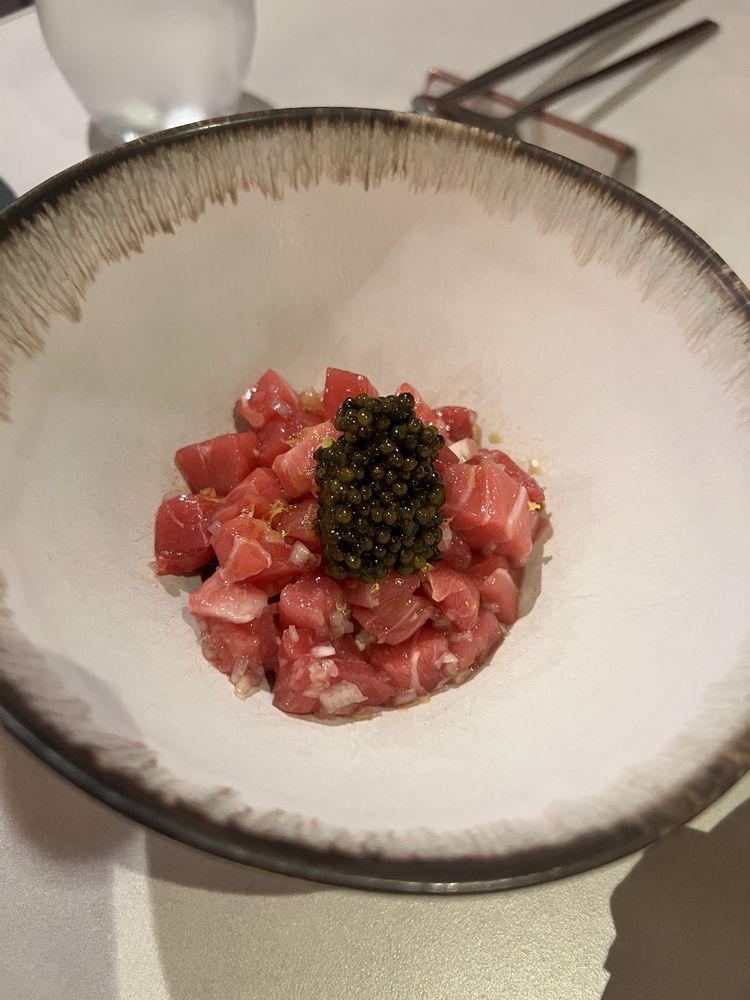 A shallow, wide ceramic bowl with a delicate brown-rimmed edge holds a small, artful mound of finely diced raw bluefin tuna, lightly dressed. Atop the tuna sits a neat dollop of glossy black caviar, creating an elegant contrast of color and texture. In the background, metal chopsticks and a water glass are visible.