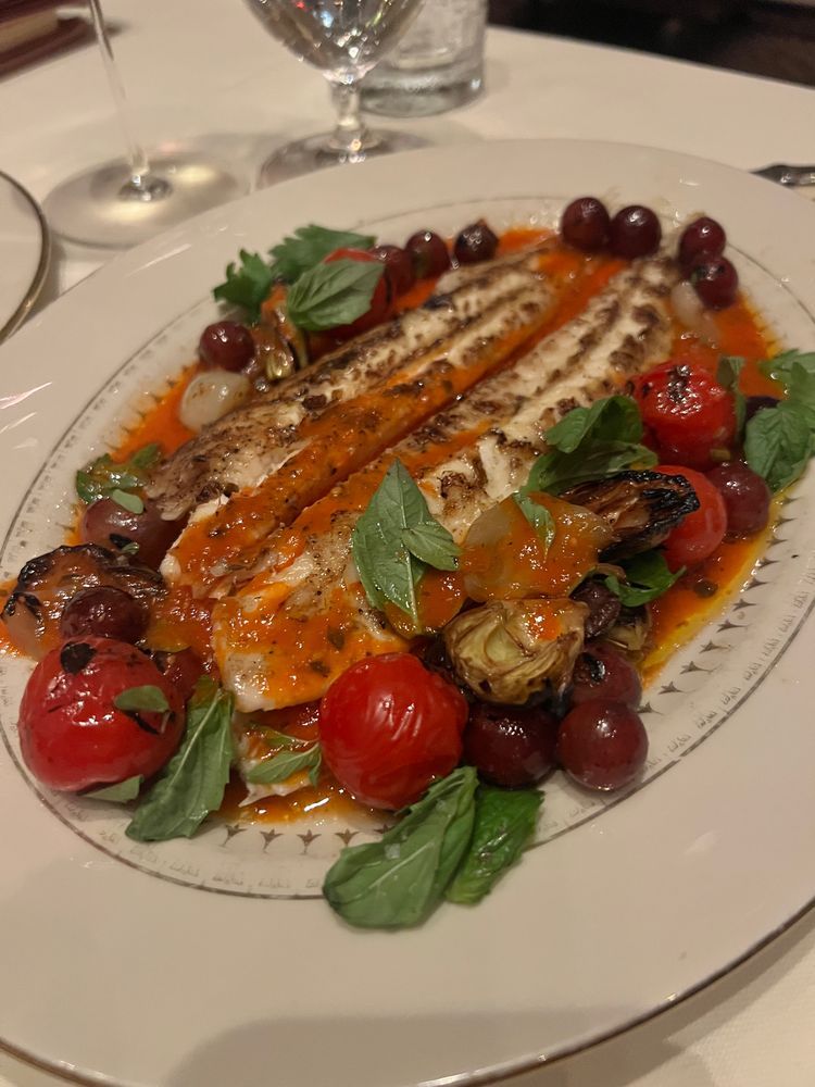 A beautifully plated fish entrée featuring two grilled fillets surrounded by a bright orange tomato-based sauce, garnished with roasted cherry tomatoes, plump grapes, artichoke hearts, and fresh basil leaves. The dish sits on elegant china, with wine glasses in the background adding to the luxe feel.