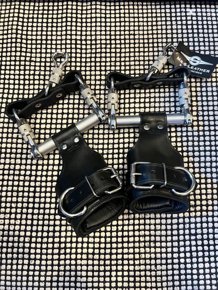 A pair of heavy-duty black leather suspension cuffs with thick metal buckles, D-rings, and aluminum handles. The cuffs are connected by metal quick links and carabiners. A branded tag from “Mr. S Leather San Francisco” is attached, all laid out on a black and white grid-patterned fabric background.
