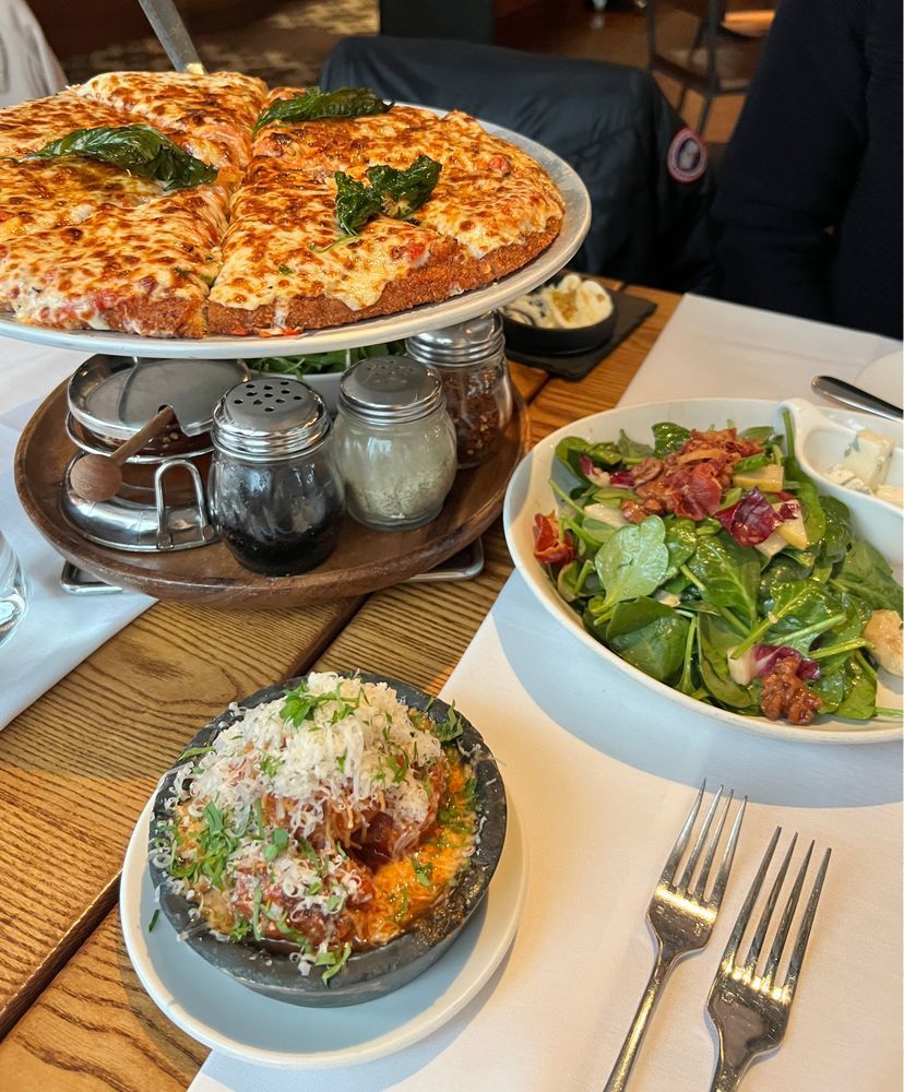 A table set for lunch featuring three dishes: a deep-dish style chicken parm pizza on a raised stand, topped generously with melted cheese and fresh basil leaves; a bowl of fresh green salad with spinach, walnuts, cheese, and bits of prosciutto; and a small black bowl filled with saucy meatballs generously topped with grated cheese and herbs. There are also condiments on the pizza stand (red pepper flakes, black pepper, grated parmesan) and two forks set on a white napkin. The setting is a cozy restaurant with wooden tables.