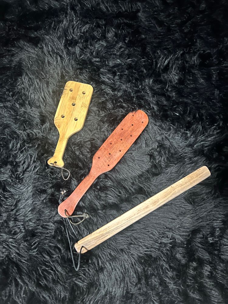 Three wooden impact toys — a light bamboo paddle with holes, a reddish paddle with a longer narrow shape and multiple small holes, and a simple straight wooden rod — are laid out on a plush black fur-like surface.