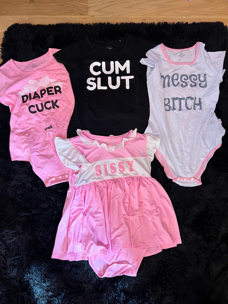 A collection of five fetish-themed adult onesies and outfits laid out on a black furry surface. From left to right, the garments read: “DIAPER CUCK” in bold black letters on a pink onesie with a white bow graphic, “CUM SLUT” in white block letters on a black shirt, “messy BITCH” in blue bubble letters on a white onesie with pink trim, a frilly pink dress-style onesie with “SISSY” in large pink letters across the chest, and all outfits appear to be styled for adult age play or sissy fetish wear.