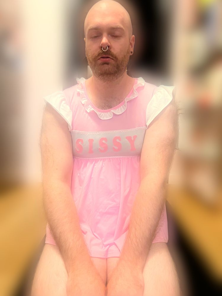 A submissive kneels on the floor with eyes closed, hands resting on their lap, wearing a soft pink outfit with the words “sissy” across. Their posture is prim and posed, knees together, conveying obedience and deliberate humiliation.