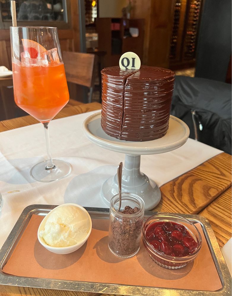 A stylish restaurant table setting featuring a decadent chocolate layer cake with glossy ganache and a white chocolate disc labeled “QI” on top, placed on a small pedestal. Beside it is a tray holding three accompaniments: a bowl of creamy vanilla ice cream, a small glass jar of chocolate crumble with a tiny spoon, and a glass bowl of glistening cherries in syrup. To the left of the cake is a tall wine glass with an orange-red cocktail, garnished with ice and a slice of citrus. The background shows wooden furniture and a warmly lit interior.