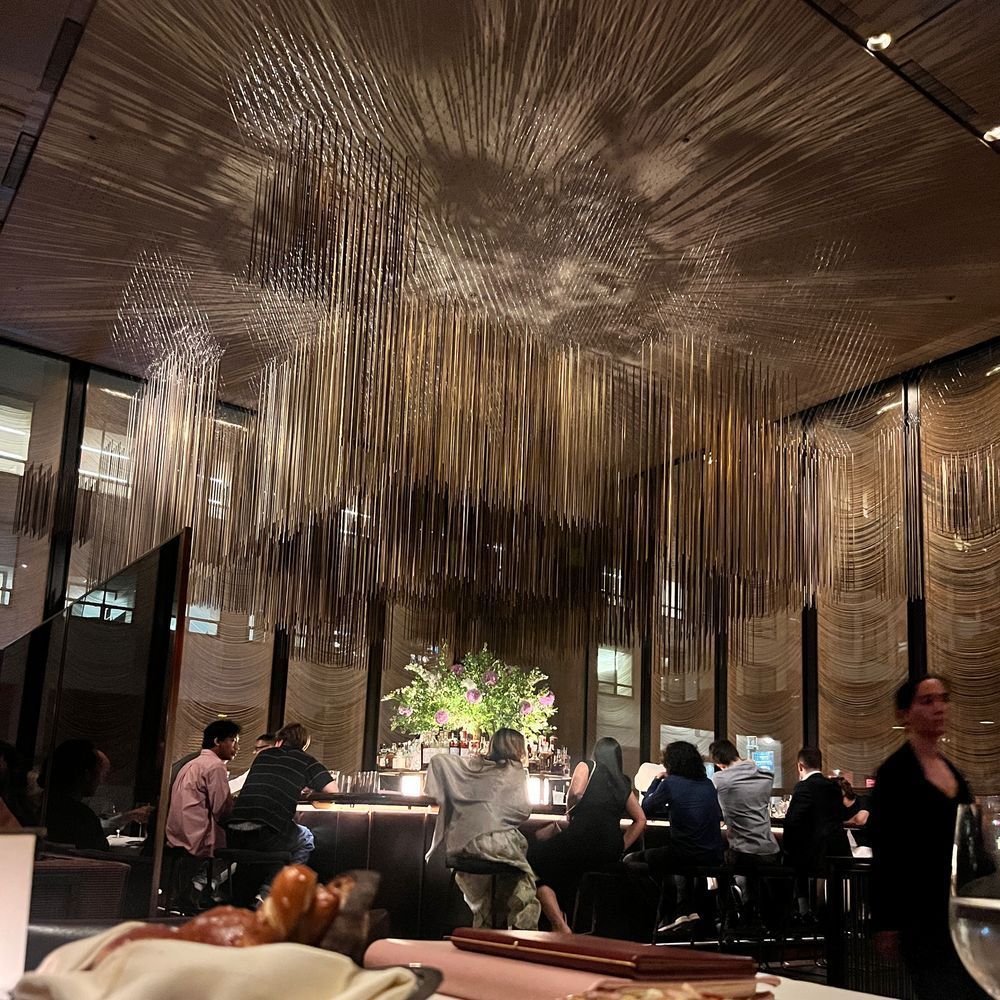 A glamorous shot of The Grill’s iconic bar area, crowned by a dazzling ceiling installation of hanging gold rods that cast intricate shadows above. The bar is surrounded by patrons enjoying cocktails, with a lush floral arrangement as the centerpiece, creating a scene of classic Manhattan opulence.
