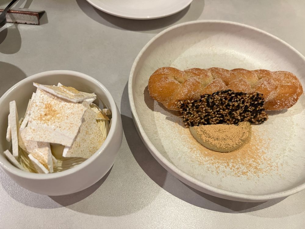 A dessert presentation with two plates. One plate holds a twisted, sugar-dusted Korean-style donut paired with a piece of sesame brittle and a dusting of nutty powder. The other dish contains a scoop of ice cream or custard topped with airy, crisp white meringue shards. Both sit on a pale table, ready to be enjoyed.