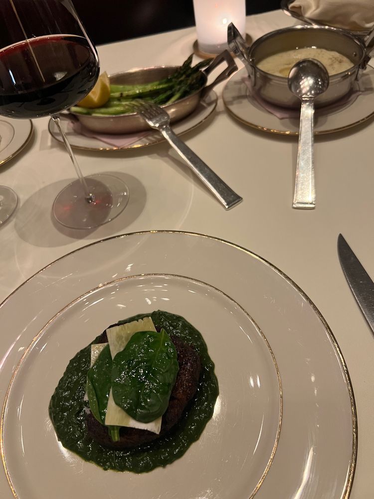 A refined dinner plate showcasing a petite filet or similar cut of beef resting on a vivid green bed of creamed spinach, topped with tender spinach leaves and thin slices of what appears to be cheese. Nearby on the table: a copper pan of roasted asparagus with lemon, a pot of velvety sauce with a ladle, and a glass of deep red wine.