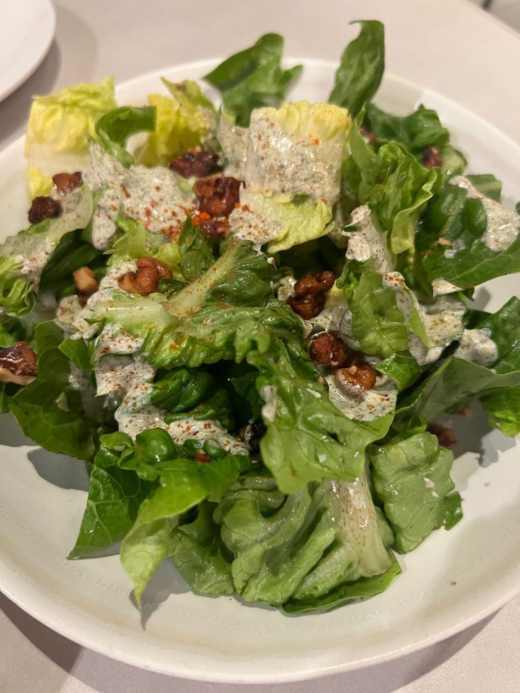 A small white plate piled with vibrant green romaine lettuce leaves, dressed with a creamy, speckled sauce. Scattered among the greens are small crunchy walnuts, lightly dusted with a reddish spice, adding texture and warmth to the fresh salad.
