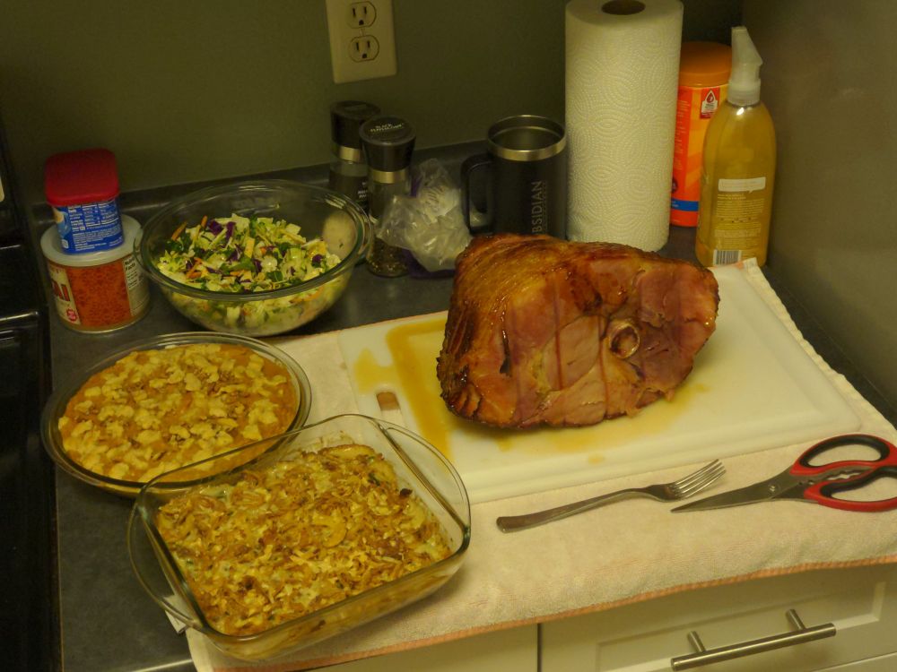 Kitchen counter with a spiral-cut ham, green beans casserole, sweet potato casserole, and bowl of salad.