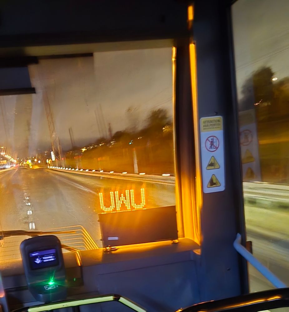 Inside view of the front of a bus travelling a highway at night, a reflected sign says "UWU".