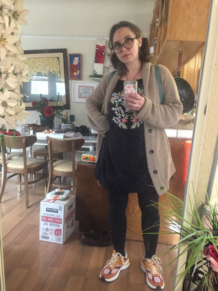 mirror selfie of white woman with slightly curly medium length brown hair in pigtails and glasses with round black frames in black tights and a black dress with pastel coral, blue, and purple-pink flowers, green leaves, and white accents embroidered on the chest in a folk art like style, with an oversized beige mens cardigan over the top, orange, pink, purple, and white running shoes, and teal backpack straps showing over her shoulders. Behind her is a kitchen counter and a messy living/dining room. The mirror is framed by fake flowers. 