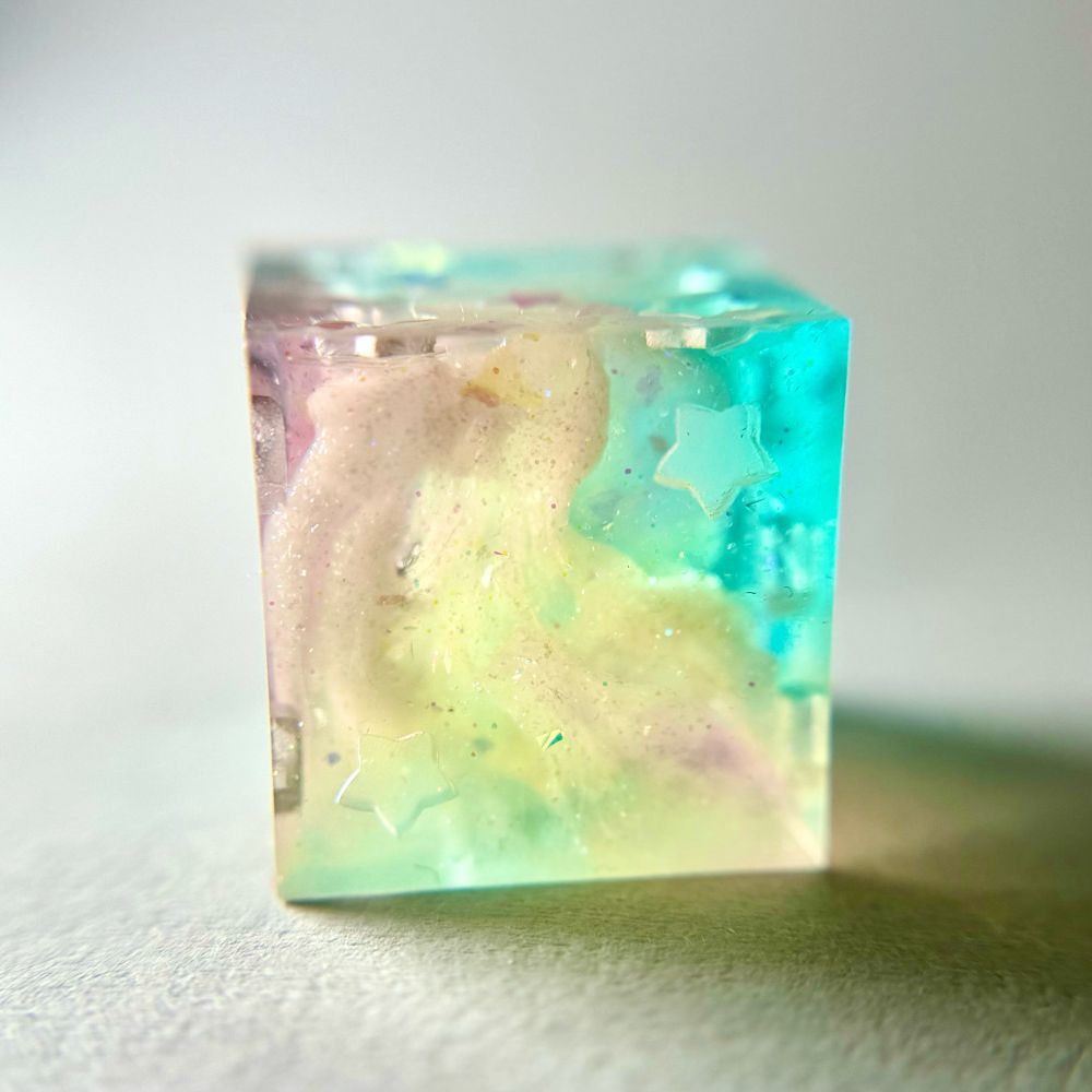 A d6 dice with star shaped pips. We are looking at the two side. The dice is translucent pink, blue, & yellow, with shimmery white clouds, is uninked, & is on a white backdrop.