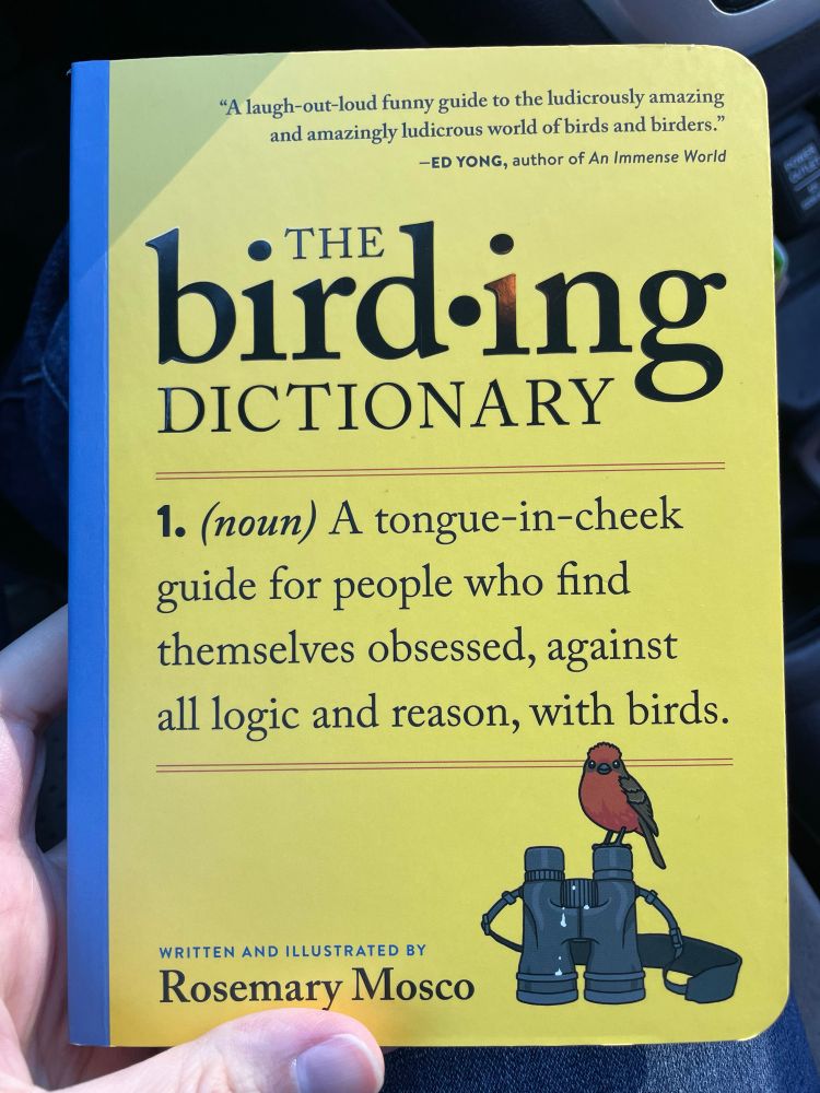 A picture of the book, “The Birding Dictonary”, by Rosemary Mosco. The cover is yellow with black text and there is a cartoon illustration of a Scarlet Flycatcher with a pair of binoculars. 