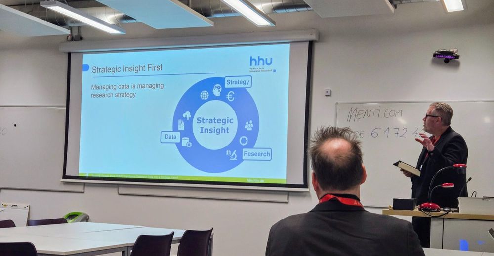 Sicht aus dem Publikum auf die Präsentation auf der Beamerleinwand und den Präsentator rechts. Gezeigt wird ein Kreis, in dessen Mittelpunkt "Strategic Insight" steht. Außenrum stehen die Begriffe "Strategy", "Research" und "Data". Es geht um "Managing data is managing research strategy".