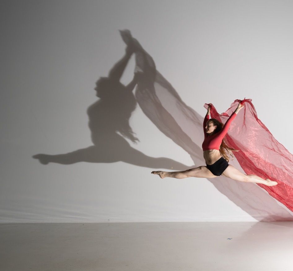 
CD: Picture of Zahna leaping in the air wearing black shorts and a long sleeve red crop top holding red fabric flowing behind her. The silhouette shadow doubles on the wall. End Description.