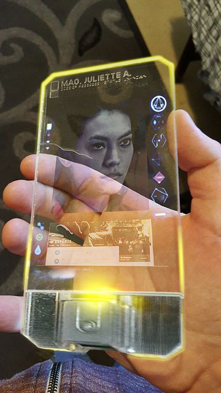 A still from the TV show The Expanse showing the right hand of a white male holding a phone-sized device. The device is almost entirely transparent and, among other widgets, displays a photo of a dark haired woman, with the text at the top: "MAO. JULIETTE A."