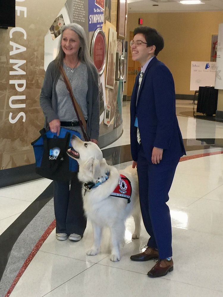 Mom is standing in a university hallway holding a bag while Sampson wearing his service dog vest is beside her looking up happily at her focused on her. There is another person wearing a blue suit beside them. Mom and the other person are smiling. 