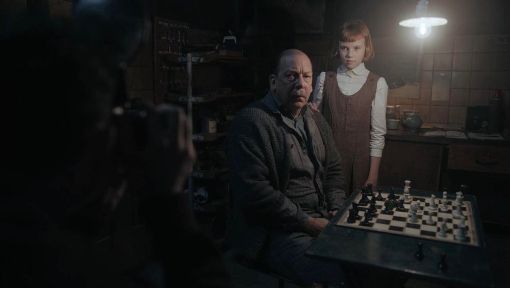 Tv photo of a stern balding man in a dark basement seated in front of a chessboard with a little redheaded girl about 10 years old dressed in very plain 1950s clothes posing for a photo with her m. From the fictional TV show about a little girl who grows up to become a chess champion despite challenging circumstances. What I love most is that the heroes are imperfect and there are no real villains in this show, just people. 