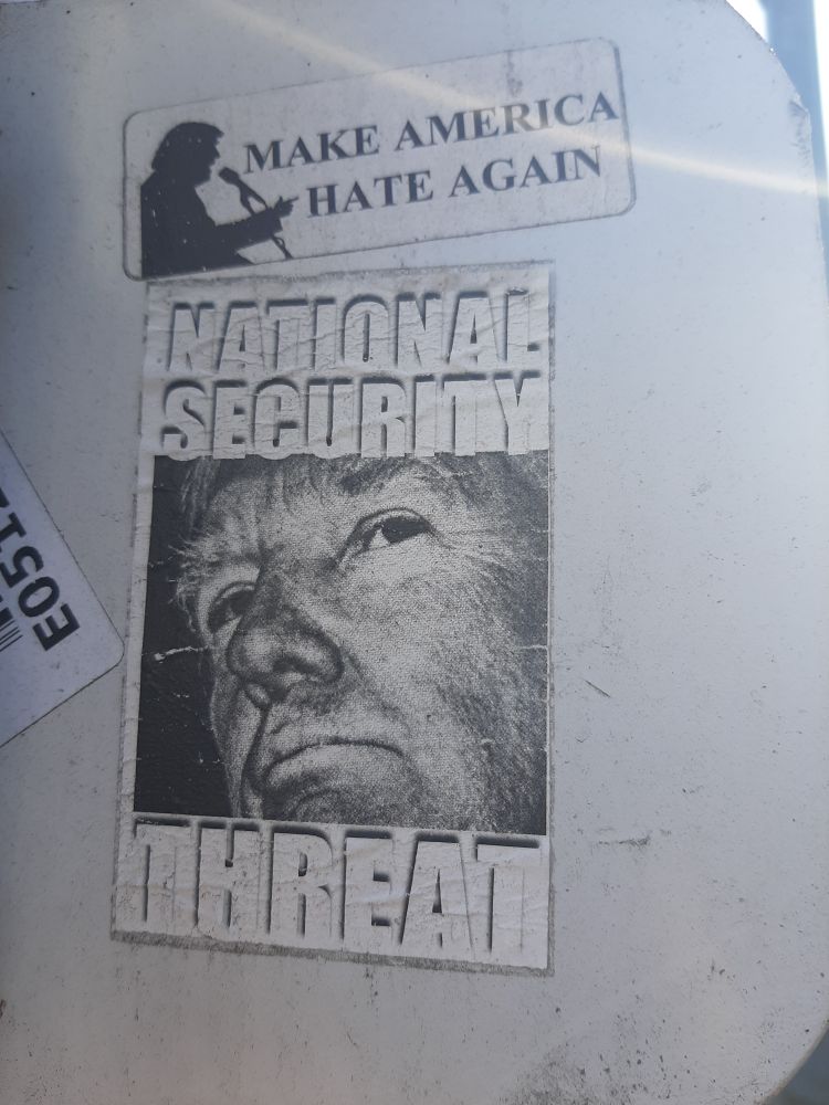 A digital photograph showing the back of a road sign, vandalized with stickers. 
A small rectangular white sticker near the top of the frame shows a silhouette of Trump at a microphone with black text reading "MAKE AMERICA HATE AGAIN"
A larger rectangular sticker shows in black & white a close up portrait Trump's old ass fugly grumpy grampa dicklord shitface with an expression of disdain. Top text in white reads "NATIONAL SECURITY" Bottom text in white reads "THREAT"