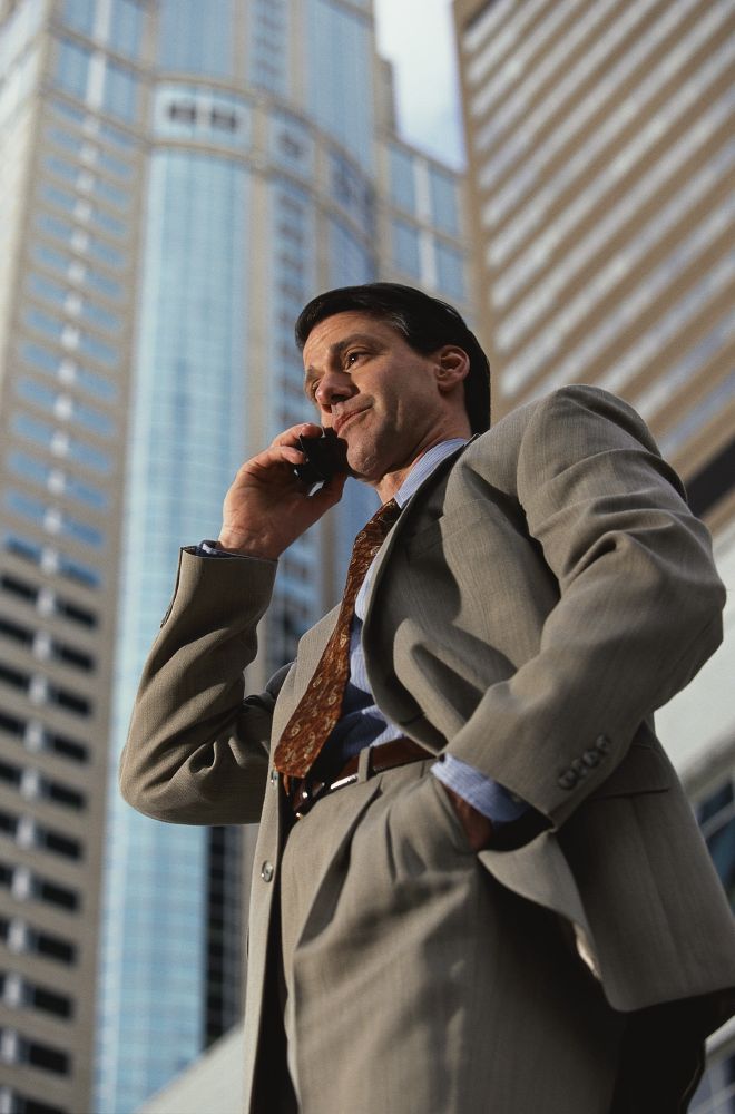 A guy in a suit on a city street doing some damn business on a cell phone. The business never stops!!!!!