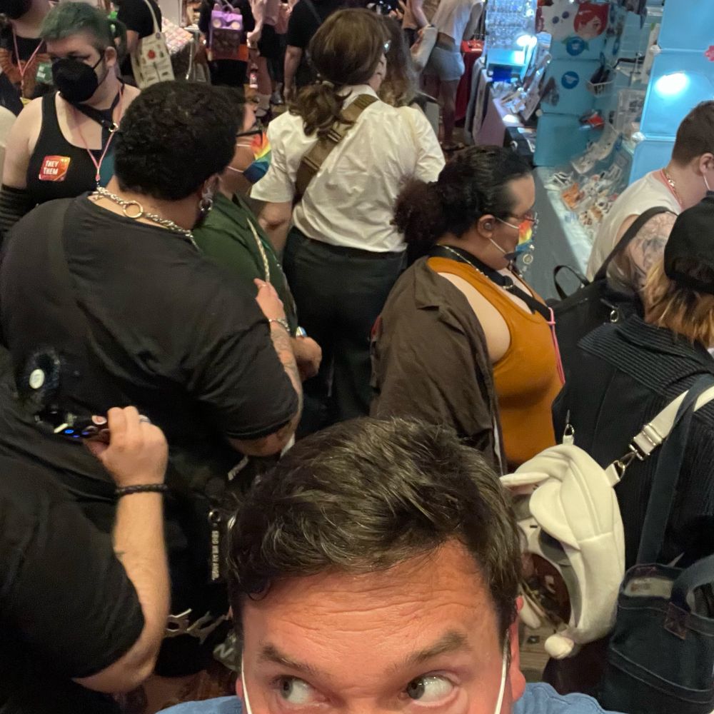 A crowded indoor scene featuring a diverse group of people, primarily seen from the perspective of someone in the foreground. Attendees are wearing masks and various clothing styles, with items displayed in the background. The atmosphere suggests a social event or gathering.