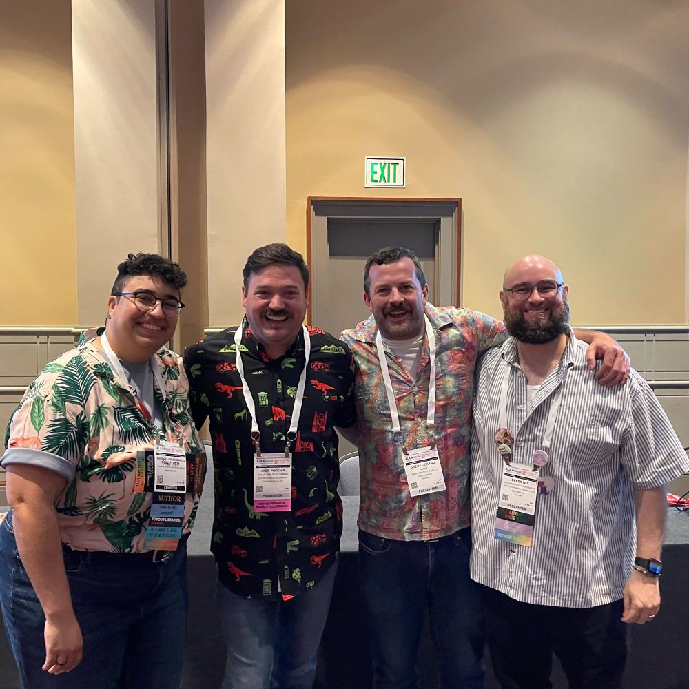 A group of four people stands together, smiling and wearing colorful, patterned shirts. They each have name badges and are posed in a welcoming indoor setting with soft lighting.
