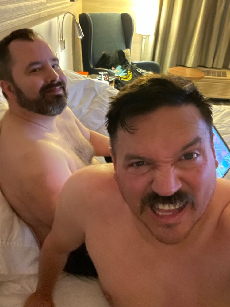 Two shirtless men are sitting on a bed in a hotel room. One man is smiling while the other is making a humorous, exaggerated face. The room has a lamp, a chair, and some bags in the background. The overall mood appears playful