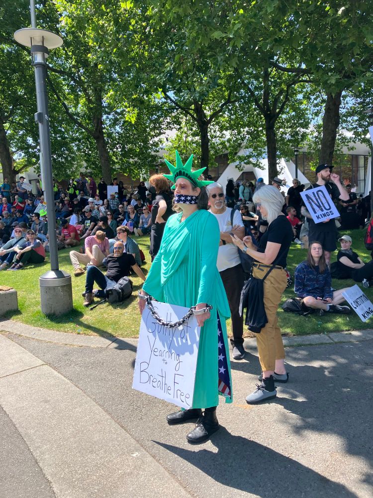 Woman dressed as Statue of Liberty gagged with American flag and hands bound holding sign saying "Guilty of yearning to be free"