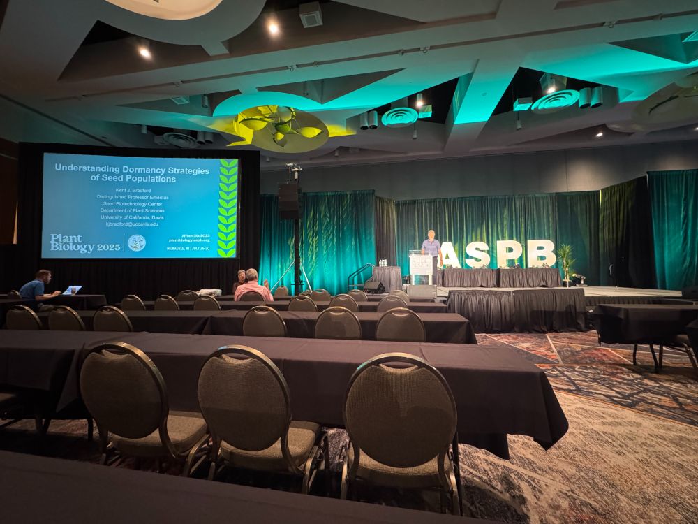 A male speaker stands on a stage with a green curtain background and big block white letters that say "ASPB" behind him. There is a lectern directly in front of him with a "Plant Biology" side across the front. On the left side of the image there is a large screen with the title page of the speaker's slideshow presentation. In the foreground of the photo there are tables lined with a black table cloth and chairs facing the speaker. 